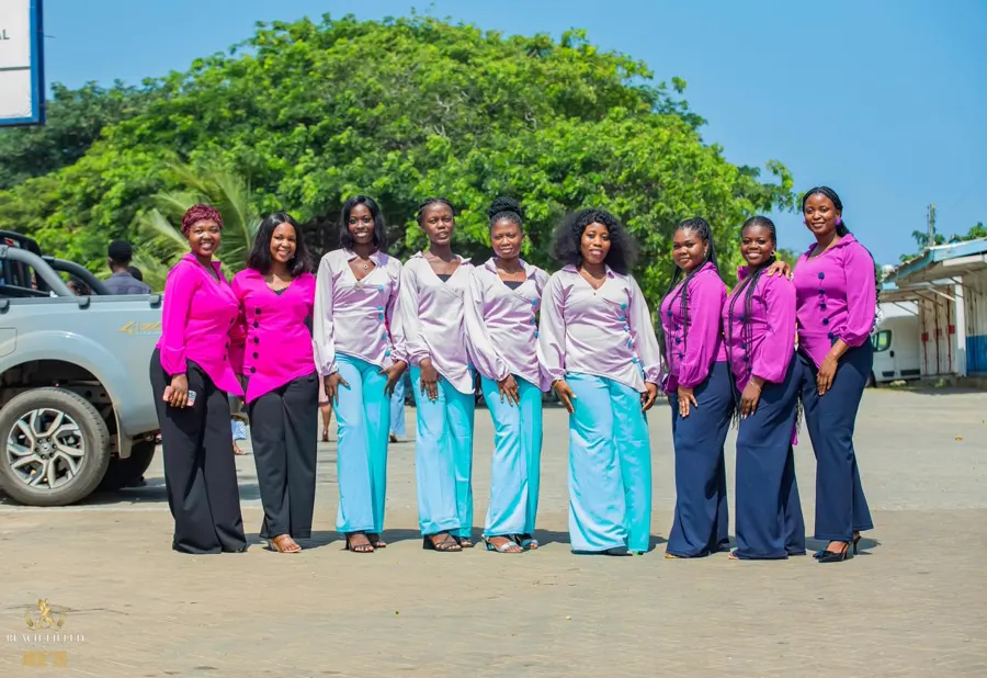 A row of women in matching pink, lilac and purple blouses smiling together outdoors