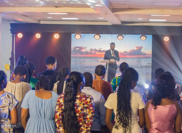 A minister in a suit speaks from a glass pulpit as the congregation stands before the stage