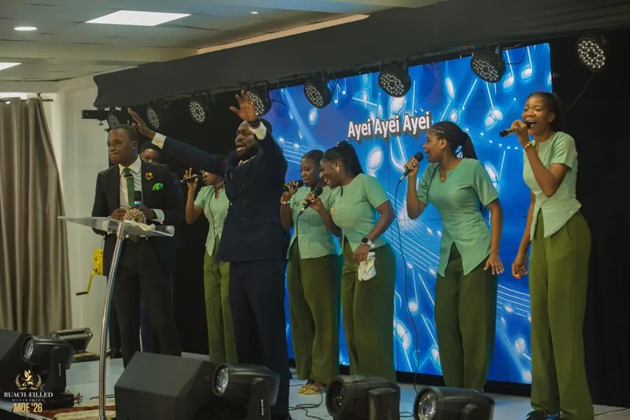 A minister with hands raised leads the worship team, dressed in matching green, in song on stage